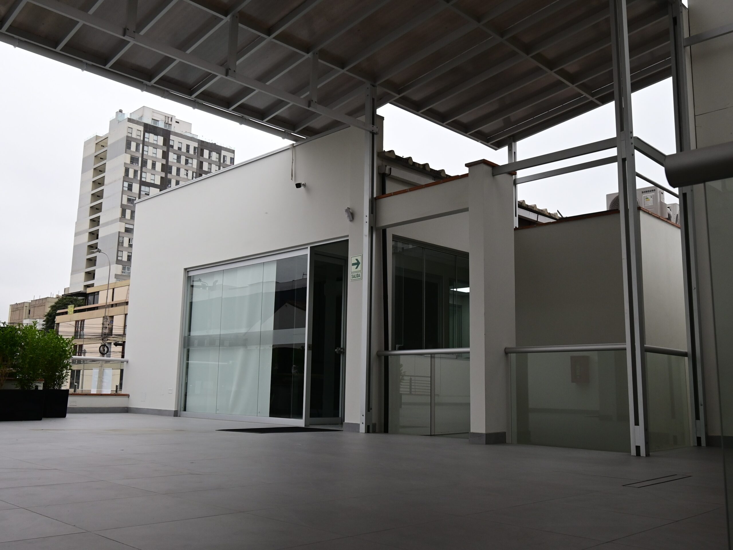 Terraza del segundo piso y acceso al área administrativa. La cobertura metálica genera un espacio exterior de uso del personal, con vistas al entorno urbano de Lince. Al fondo, el contexto residencial y comercial del distrito evidencia la inserción del centro en un tejido urbano consolidado.