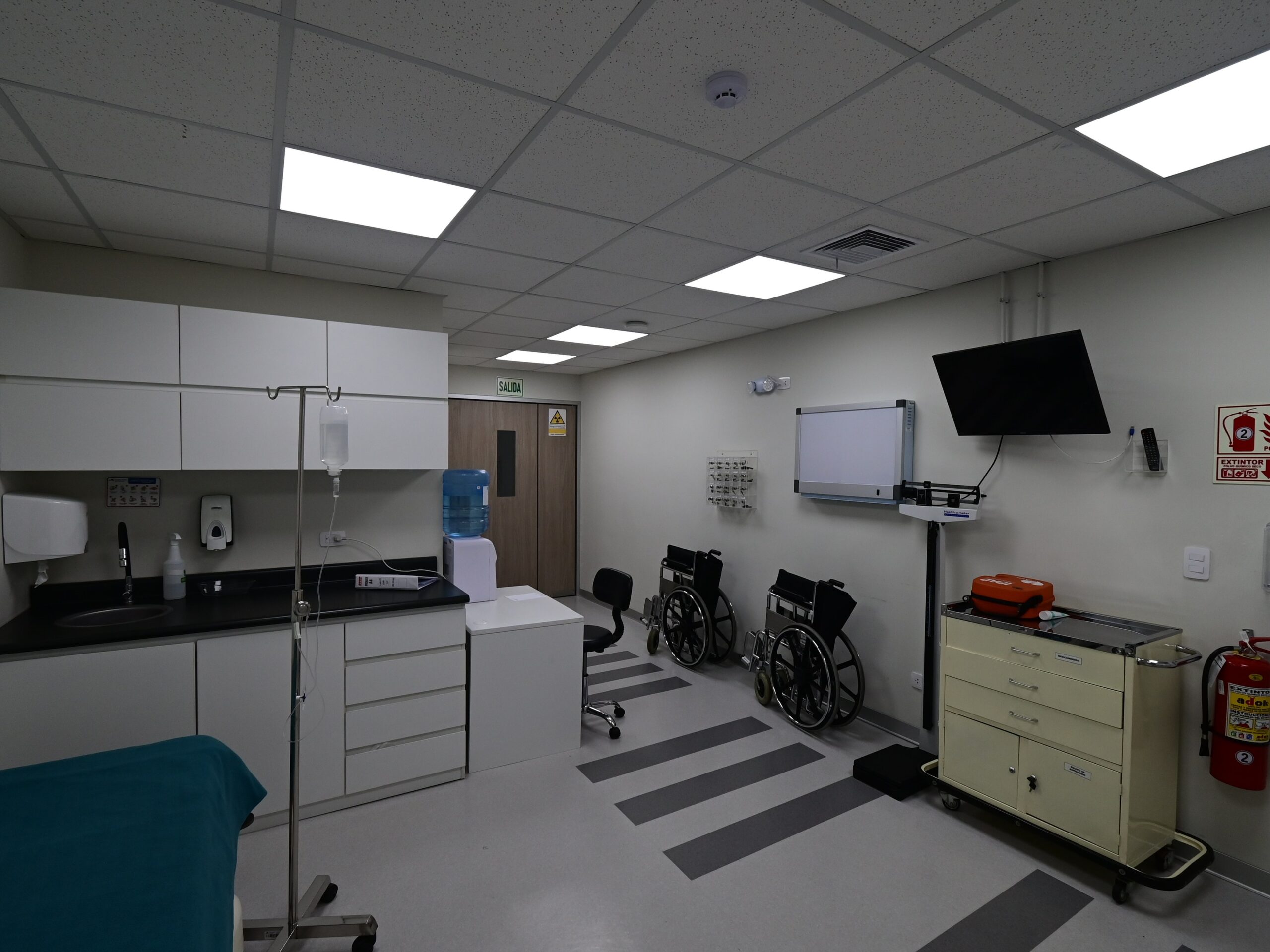 Sala de preparación del paciente. El ambiente concentra en superficie compacta el equipamiento clínico necesario para la atención pretratamiento: sillas de ruedas, carro de emergencias, monitor de signos vitales y estación de trabajo. Al fondo, la puerta de acceso al área de radioterapia con señalética de zona controlada.