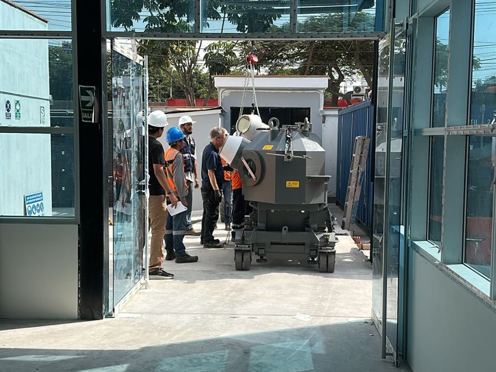 Vista desde el interior del edificio hacia el estacionamiento, donde el robot de instalación es preparado antes de ingresar al búnker. La apertura vidriada del ingreso y el equipo en primer plano ilustran la secuencia de traslado interior.
