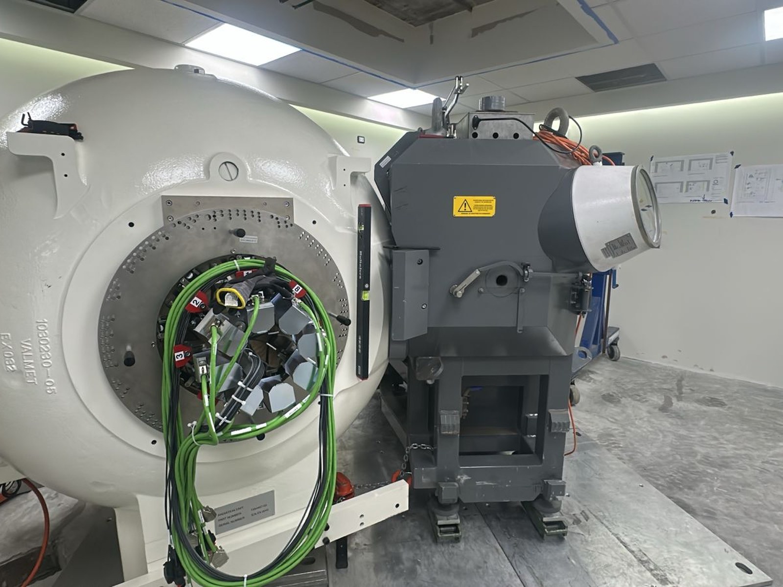 Estado avanzado del montaje: ambos componentes principales del Gamma Knife Esprit conectados dentro del búnker. Los planos del fabricante adheridos a la pared al fondo corresponden a los protocolos de instalación del equipo de Elekta.