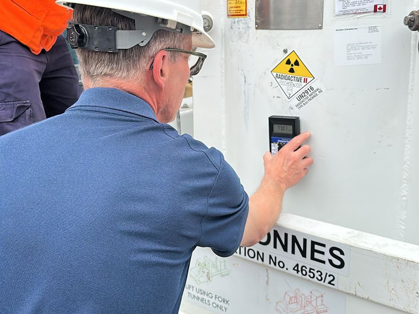 Un técnico especializado de Elekta midiendo niveles de radiación sobre el contenedor de fuentes con dosímetro, previa apertura. La señalética RADIOACTIVE II y el código de transporte UN2916 identifican el contenido. Esta verificación es obligatoria por protocolo IPEN antes de cualquier manipulación.