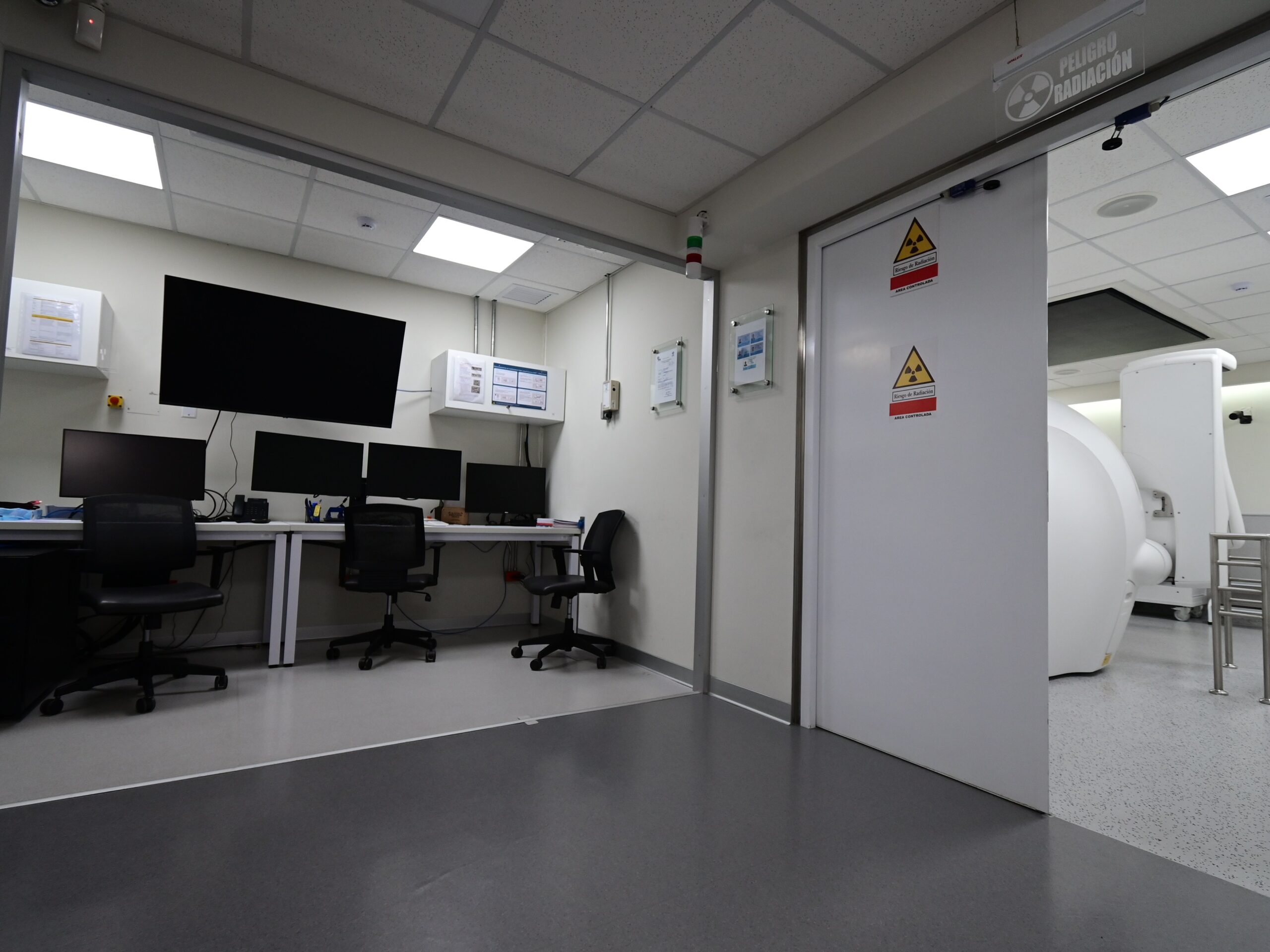 Vista desde el área de radioterapia: sala de control con estación de trabajo multipantalla; al fondo a la derecha, el equipo Gamma Knife Esprit visible a través de la puerta blindada de acceso al búnker. La señalética de zona controlada delimita el umbral entre el espacio operativo del personal técnico y el área de tratamiento.