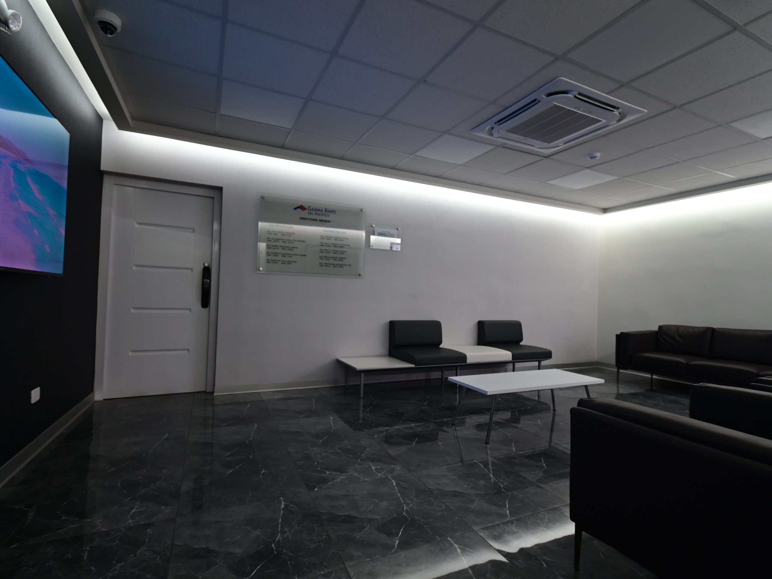 Sala de espera del primer piso. La puerta con cerradura electrónica al fondo controla el acceso al área de radioterapia; el directorio médico y los acabados de la sala comunican el nivel especializado del centro. La climatización por cassette de techo es visible en la parte superior.