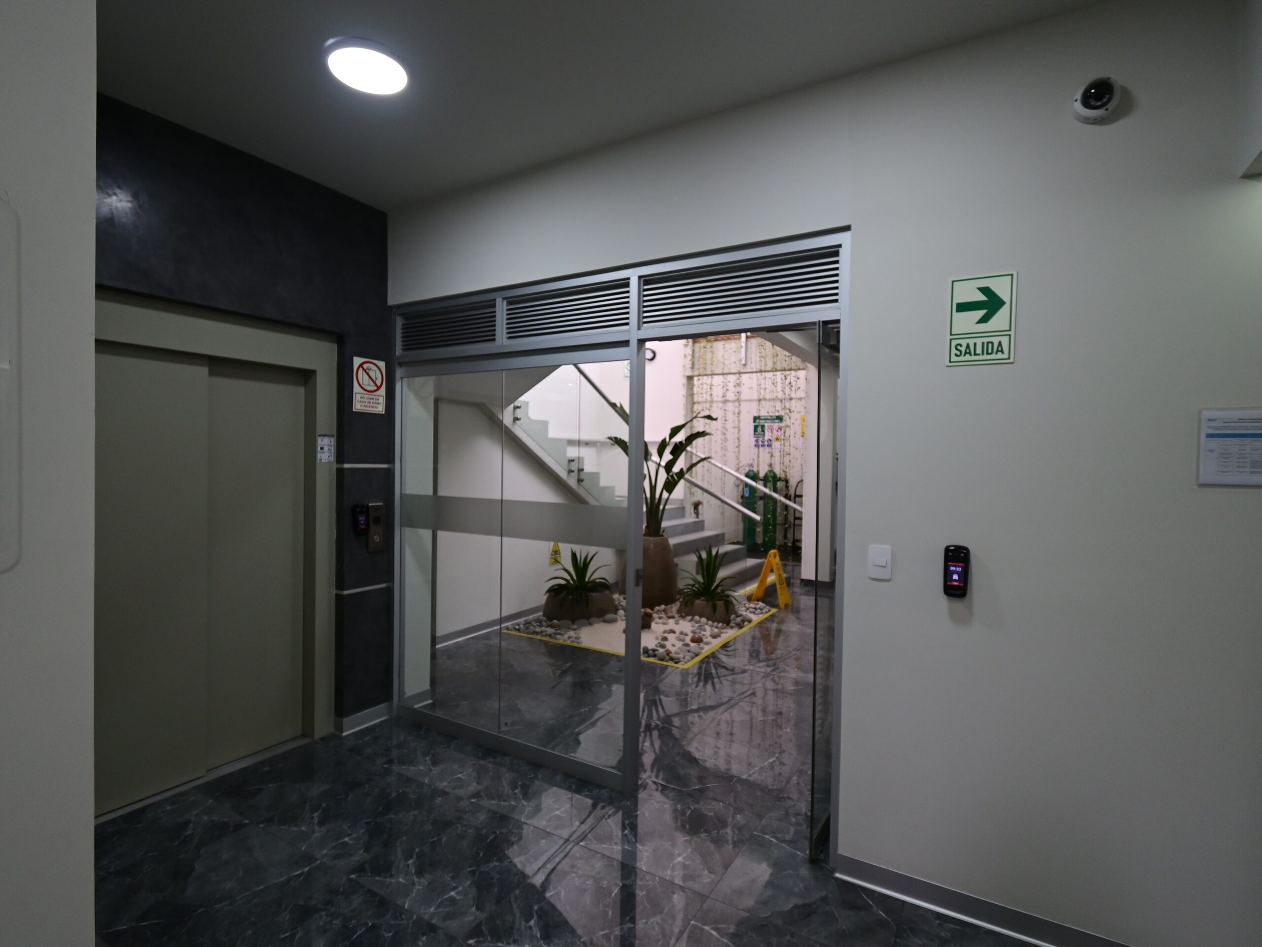 Hall de distribución hacia la zona técnica y administrativa. El control biométrico en la pared derecha regula el acceso al área privada que alberga la sala de planificación, vestidores médicos y escalera hacia las oficinas y sala de reuniones del segundo piso. La puerta vidriada separa el circuito de pacientes del circuito exclusivo de personal.