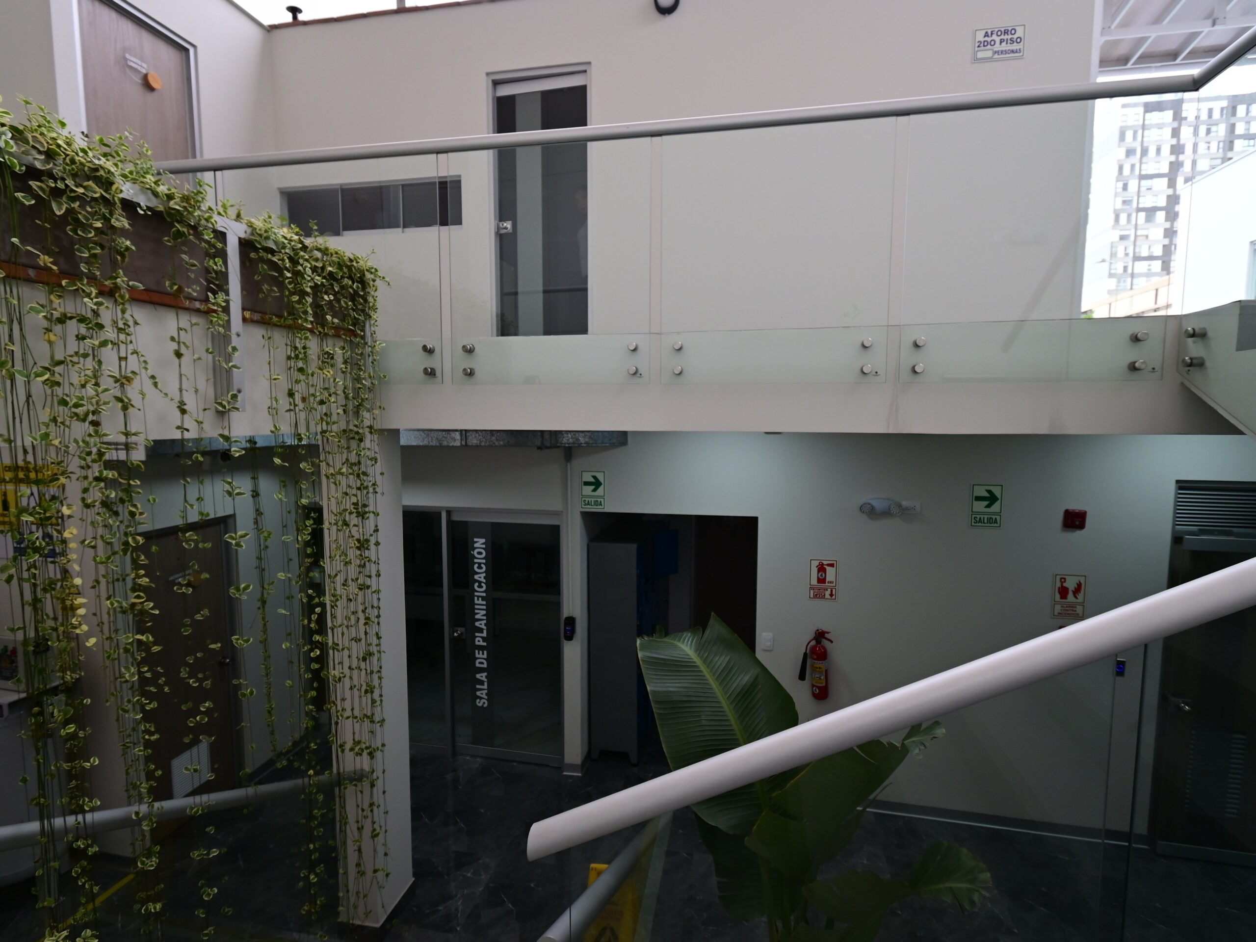 Vista desde la escalera hacia el espacio interior de doble altura. El muro verde vertical articula la relación entre el primer y segundo piso; al frente, el acceso a la sala de planificación. La condición de luz natural cenital y la presencia de vegetación interior responden a criterios de bienestar del personal técnico y médico.